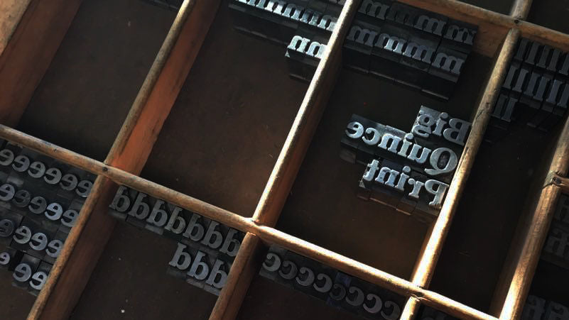 Close-up of a vintage wooden letterpress printing box with compartments holding metal type letters. Some are sorted by letter, but in a shaft of sunlight are letters spelling "Big Quince Print".